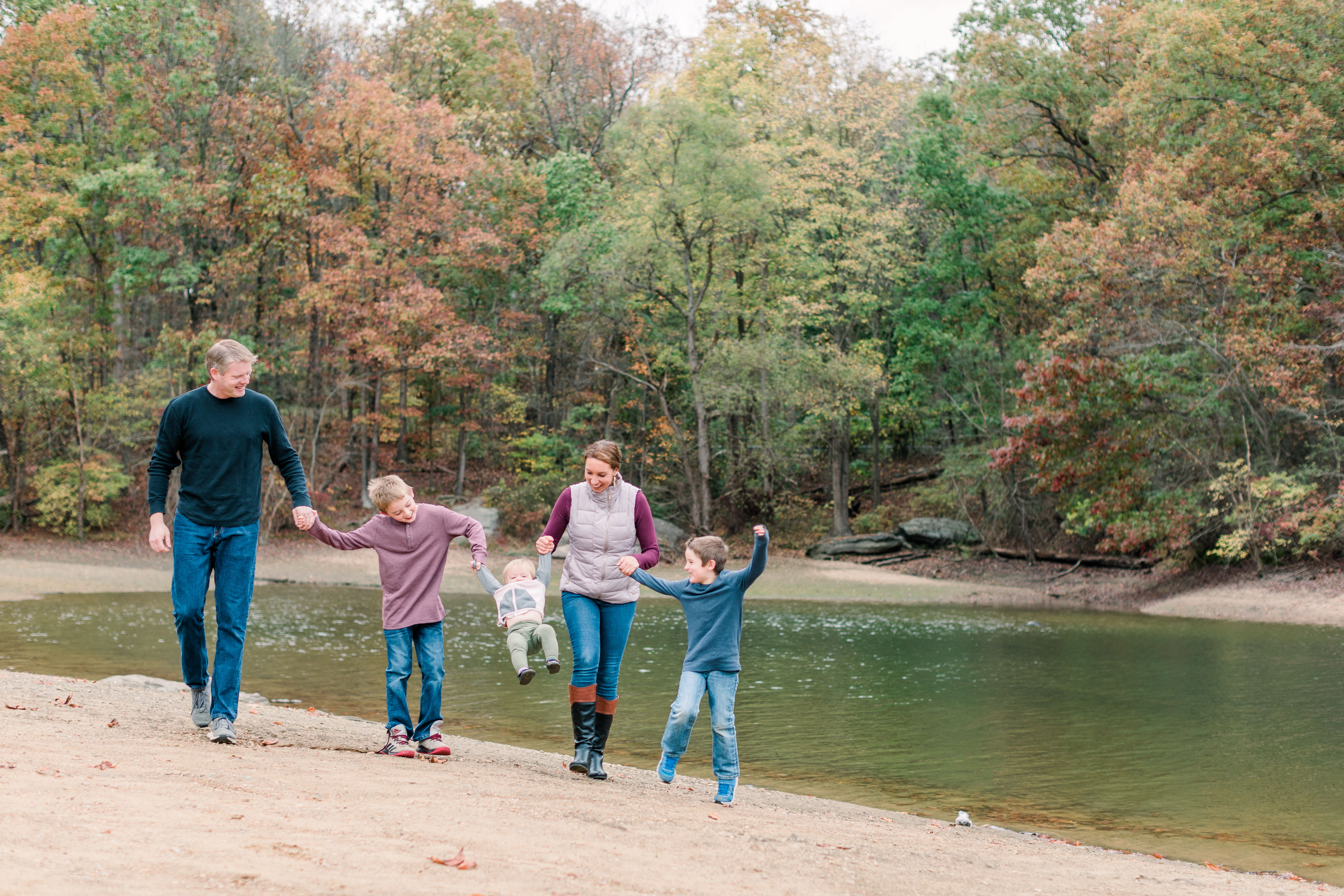 A family of 5 holds hands next to a lake with fall leaves around them. They swing their youngest.