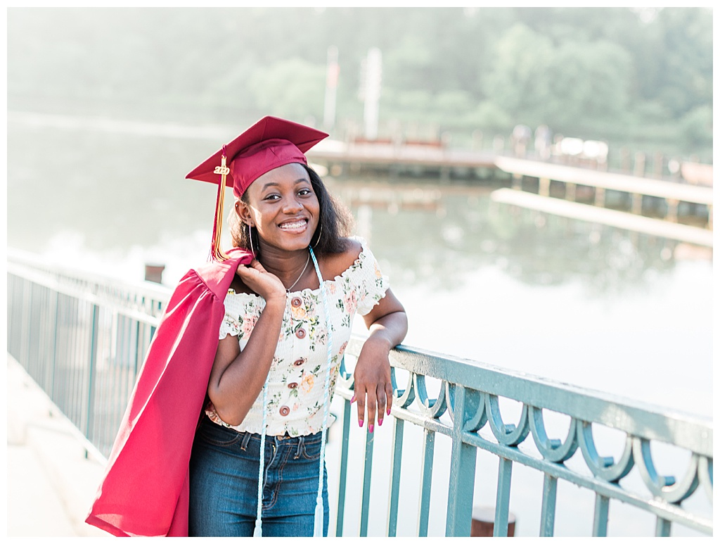 African American teenage girl leans against a rail with her graduation gown draped over her shoulder and graduation cap on her head.