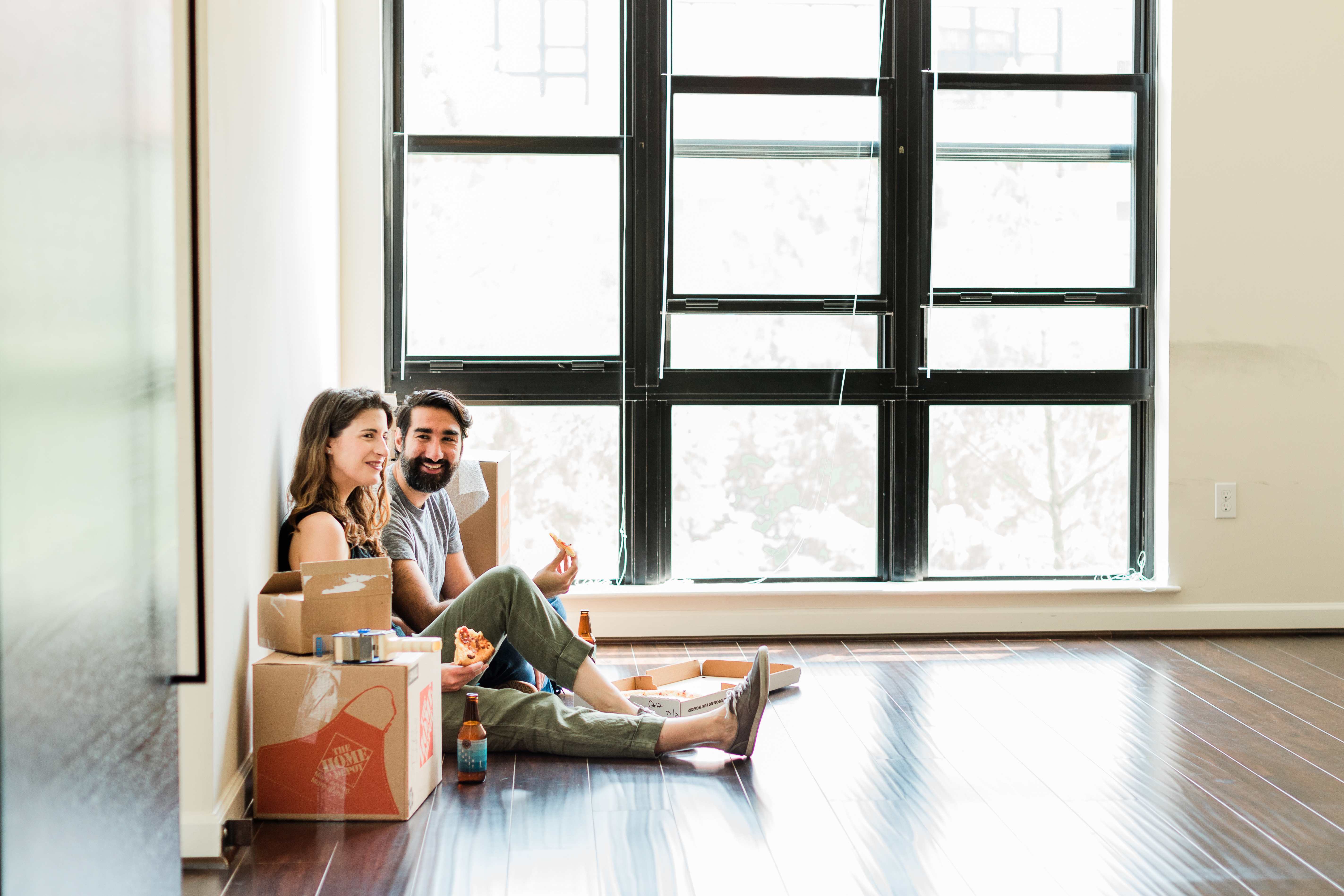 A young couple sits on the floor of their empty apartment surrounded by a few boxes. They are eating pizza, drinking beer, and laughing.