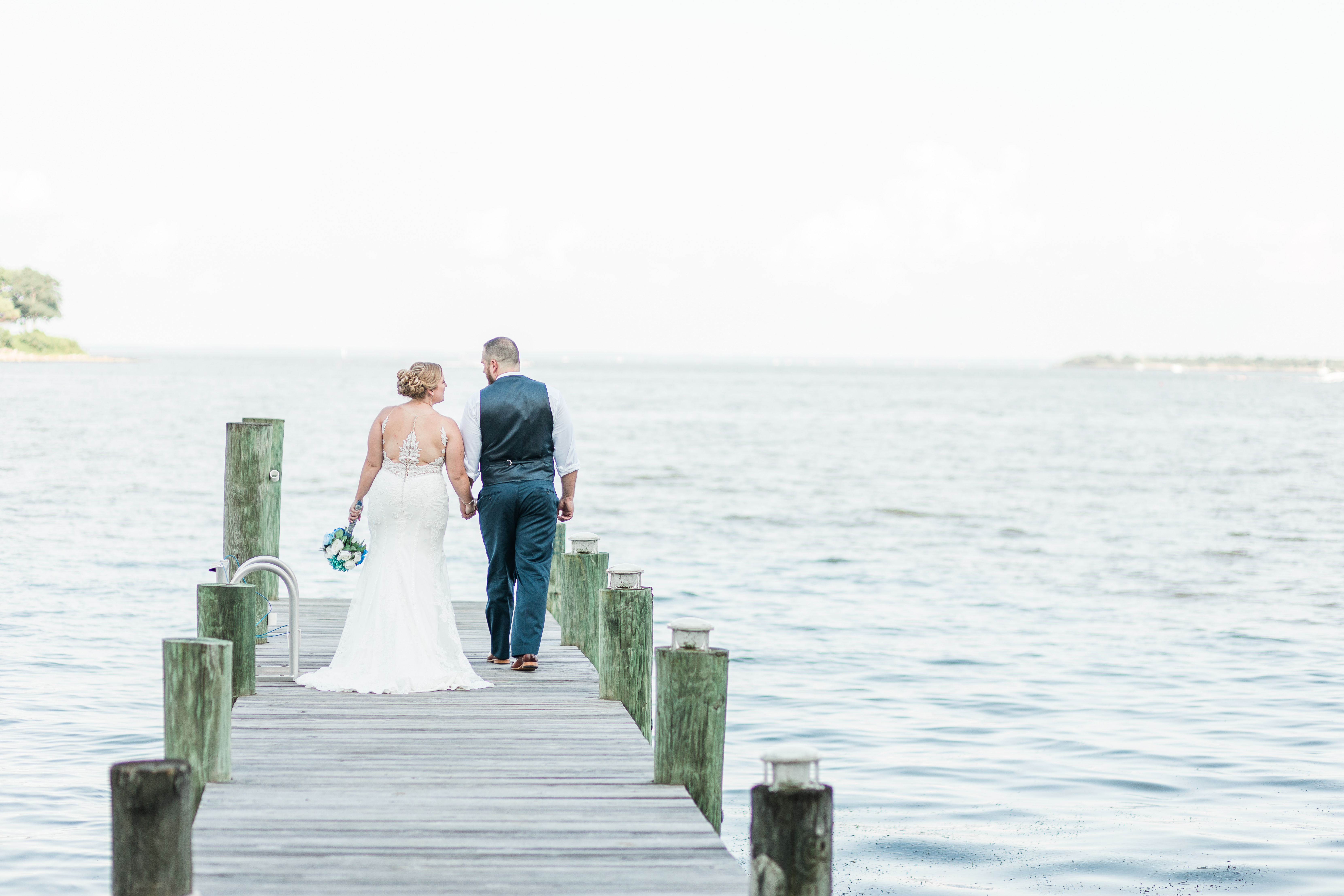 A wedding couple walk hand in hand down a dock, toward the bay