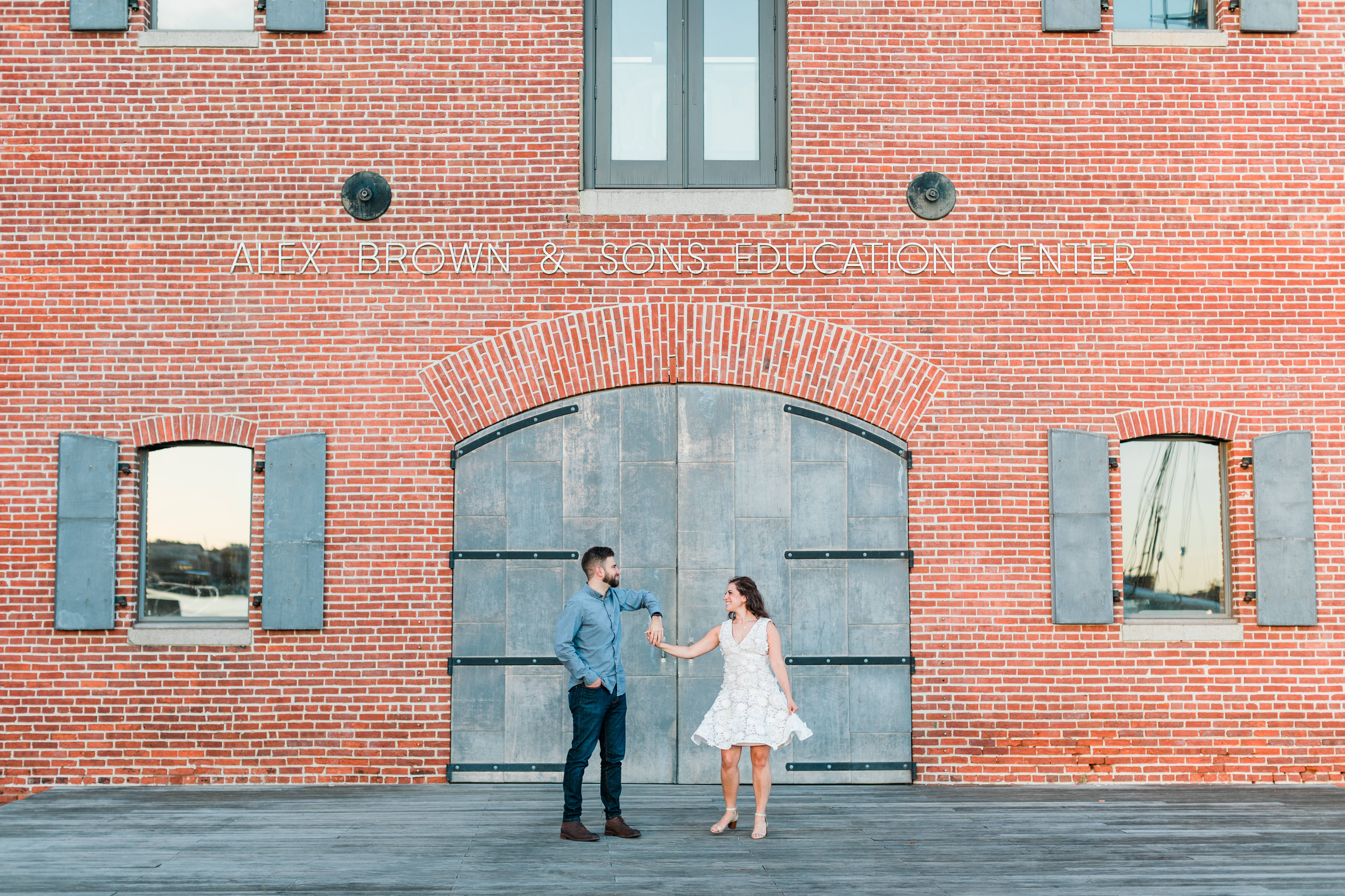A woman in a white dress and man in a blue shirt dance in front of a red brick building with blue doors in Fells Point Baltimore