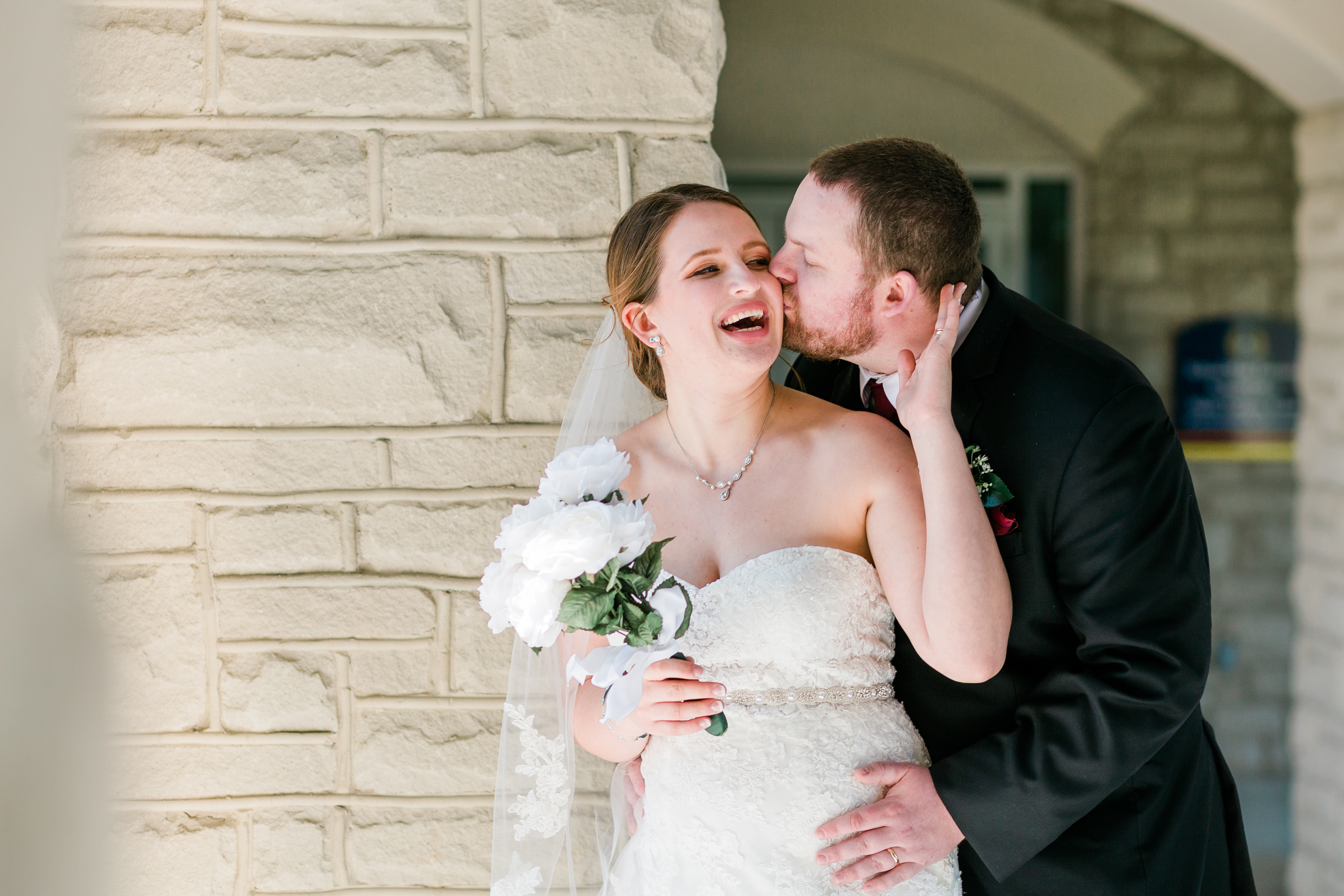 A bride laughs while her groom kisses her cheek