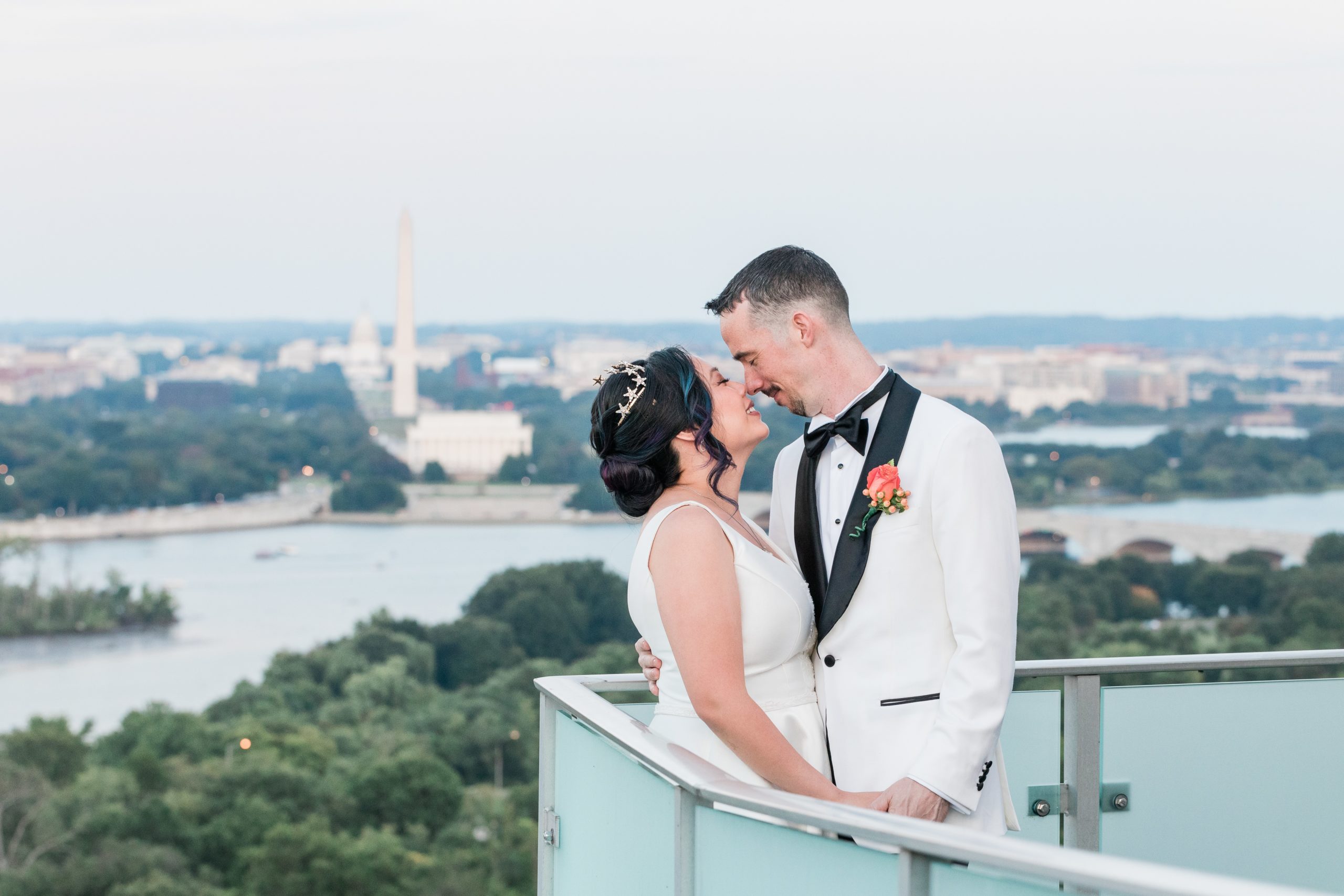 A groom in a white tuxedo jacket kisses his bride who is in a white satin ballgown. They are on a rooftop looking over the Potomac River, Lincoln memorial, Washington Monument and Capitol Building
