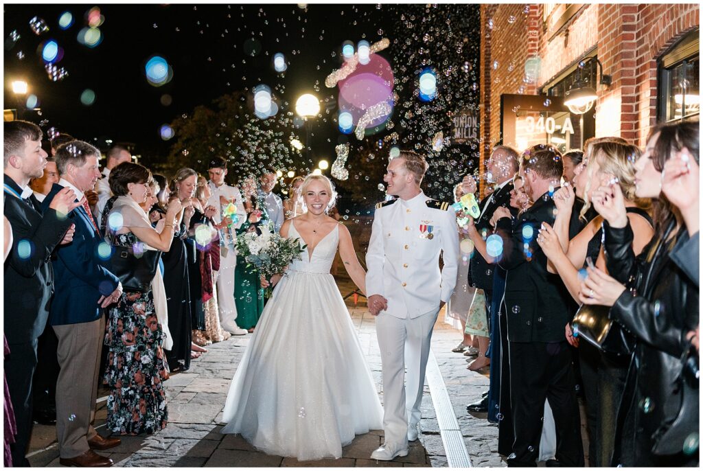 Naval officer in dress whites and bride in ballgown share joyful moment during bubble exit sendoff. Wedding guests line stone walkway blowing bubbles as newlyweds celebrate under evening lights