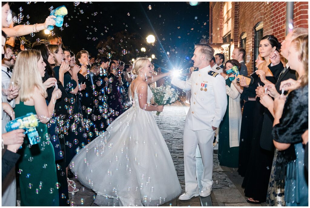 Candid moment during bubble exit as bride laughs with naval officer groom. Evening wedding sendoff with guests creating magical atmosphere with hundreds of bubbles floating in night air