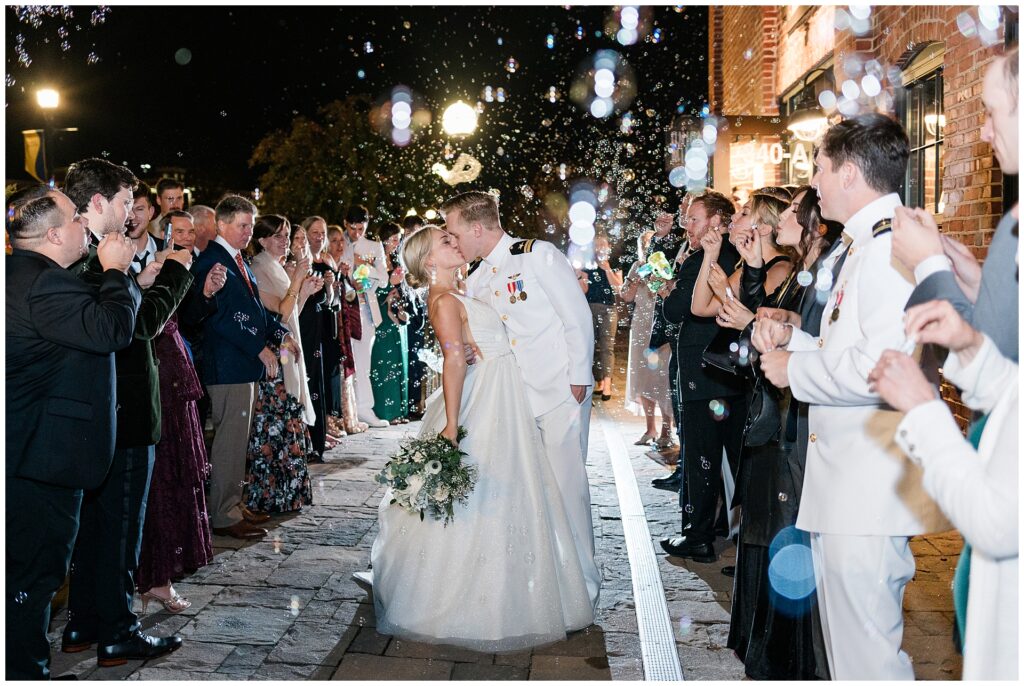 Wedding guests create magical bubble sendoff for naval officer and bride. Evening celebration captured with glowing lights and joyful guests lining stone pathway.