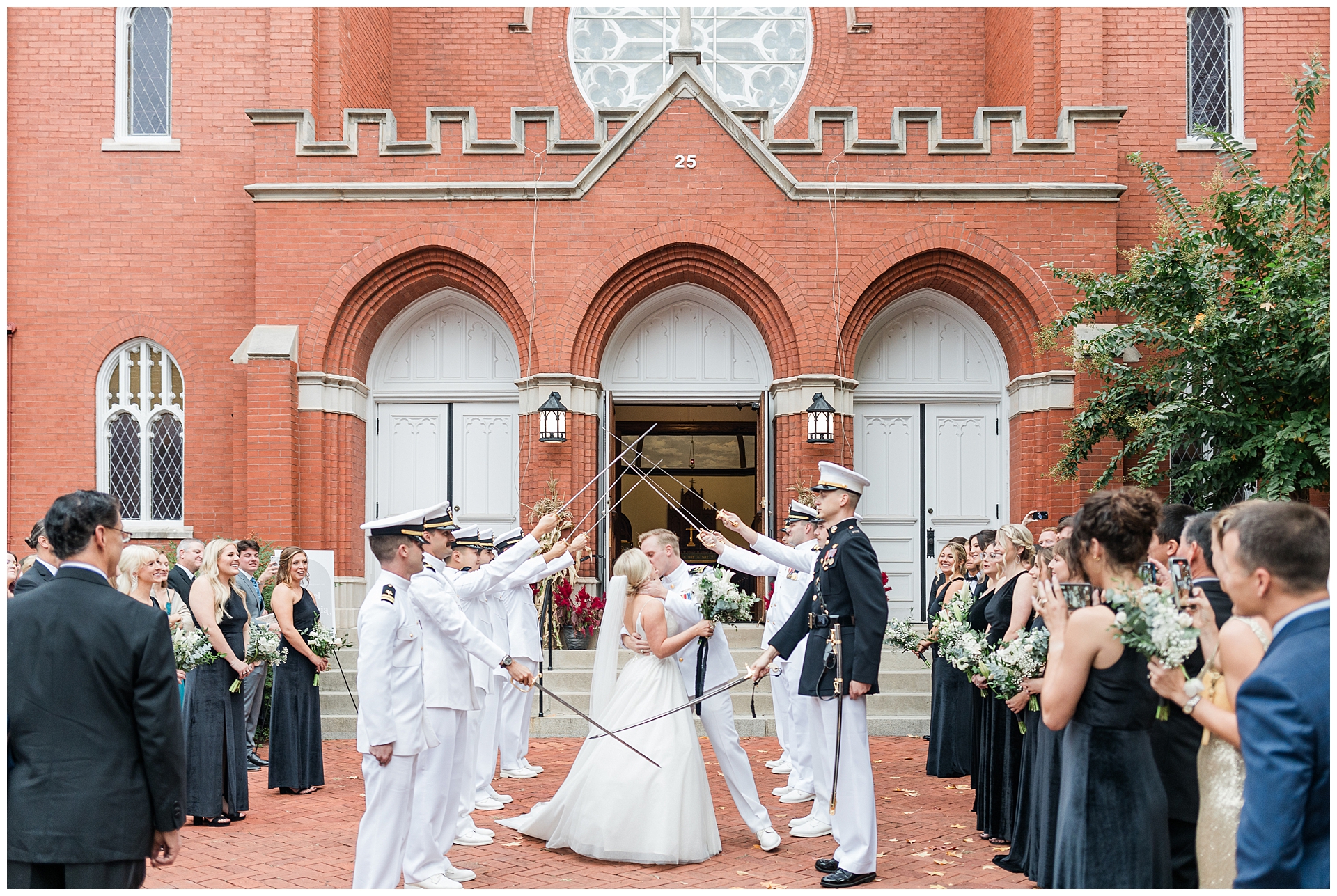 Naval officers performing traditional sword arch ceremony for newlyweds outside historic Grace United Church of Christ in downtown Frederick, Maryland. Bride in white gown and groom in naval dress whites kiss while passing through arch of swords, surrounded by wedding party in black dresses