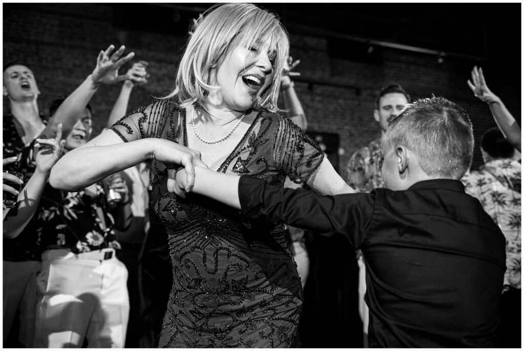 Black and white capture of wedding guest dancing with young attendee at reception. Intimate moment showing multi-generational celebration at elegant venue