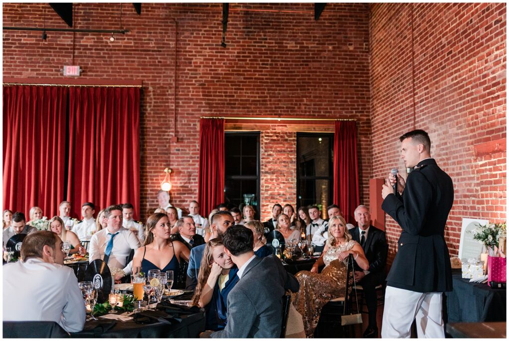 Military officer delivers wedding toast at Union Mills Public House in Frederick, MD, speaking to gathered guests including fellow service members in dress uniforms. Historic brick walls and red velvet curtains frame elegant military wedding reception.