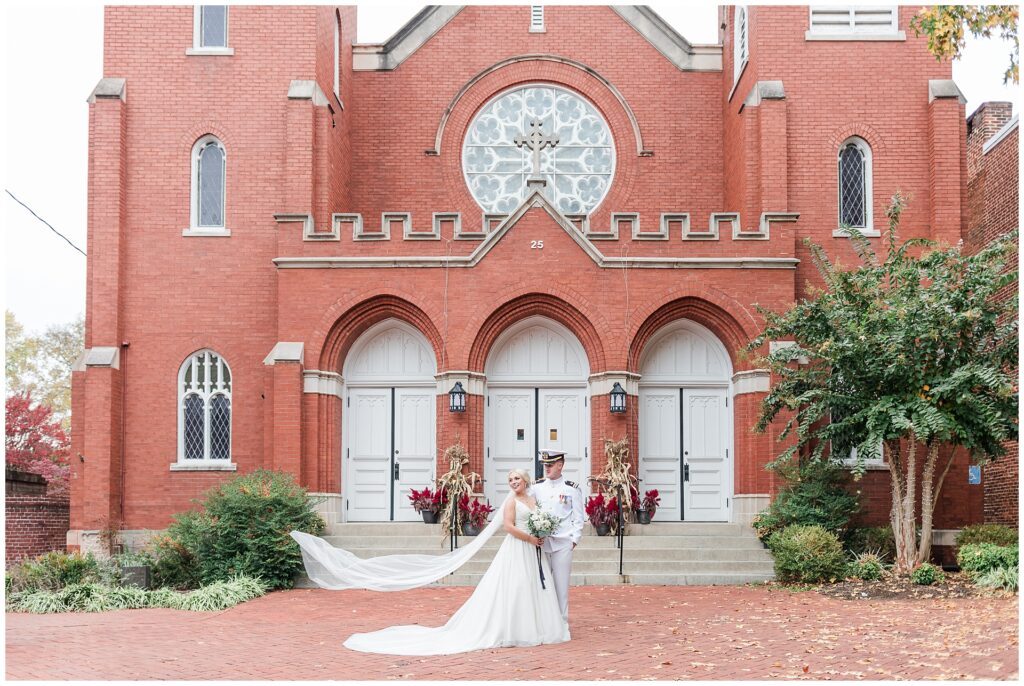 Navy officer and bride with cathedral-length veil in front of historic brick church in downtown Frederick, fall wedding portrait