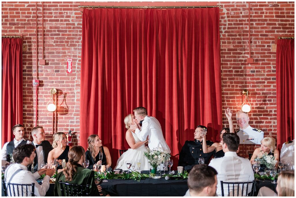 Naval officer and bride share romantic kiss during wedding toasts at Union Mills Public House, surrounded by military guests and wedding party. Elegant reception features historic brick walls, red velvet curtains, and classic black and white table settings in downtown Frederick