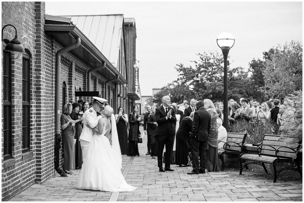 Naval officer and bride share first kiss outside historic Union Mills Public House in downtown Frederick, black and white candid moment with wedding guests