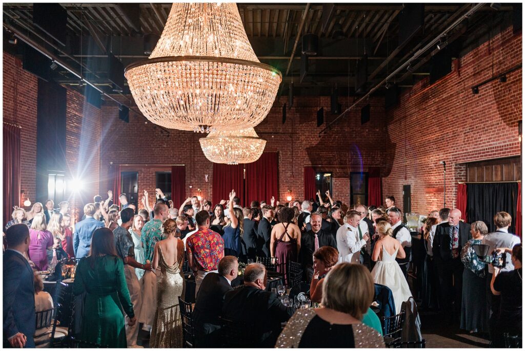 Packed dance floor at wedding reception under crystal chandelier. Guests dancing in mix of formal wear and Hawaiian shirts, showcasing the transition from black tie ceremony to lively celebration