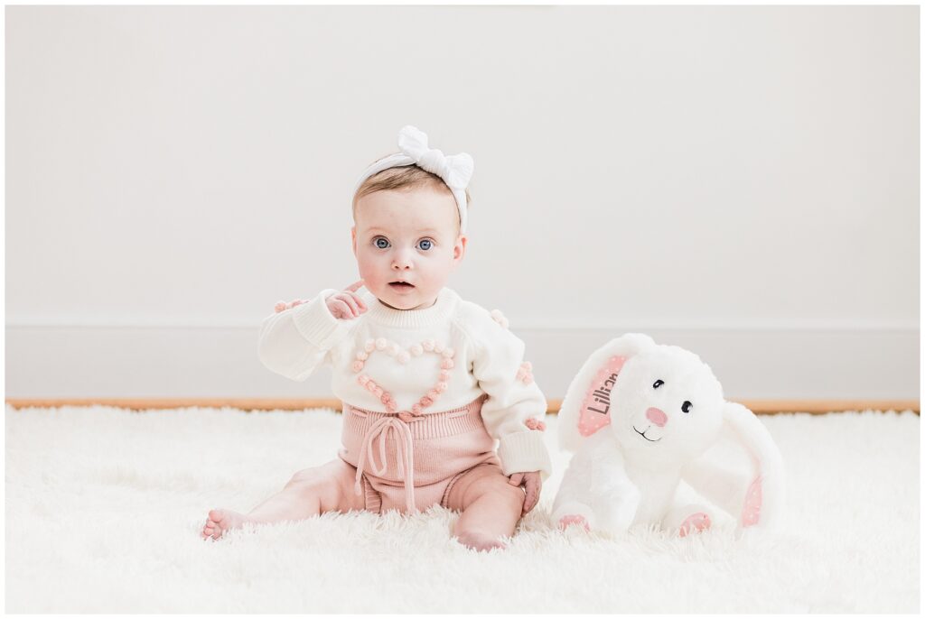 Six-month-old baby girl sitting up during milestone photo session in Maryland, wearing blush pink and ivory sweater outfit with personalized plush bunny, styled for a lifestyle portrait.