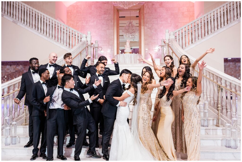 Large wedding party celebrating bride and groom kiss on marble staircase at Bellevue Conference & Event Center, showcasing full-day photography coverage at a high-capacity Virginia venue
