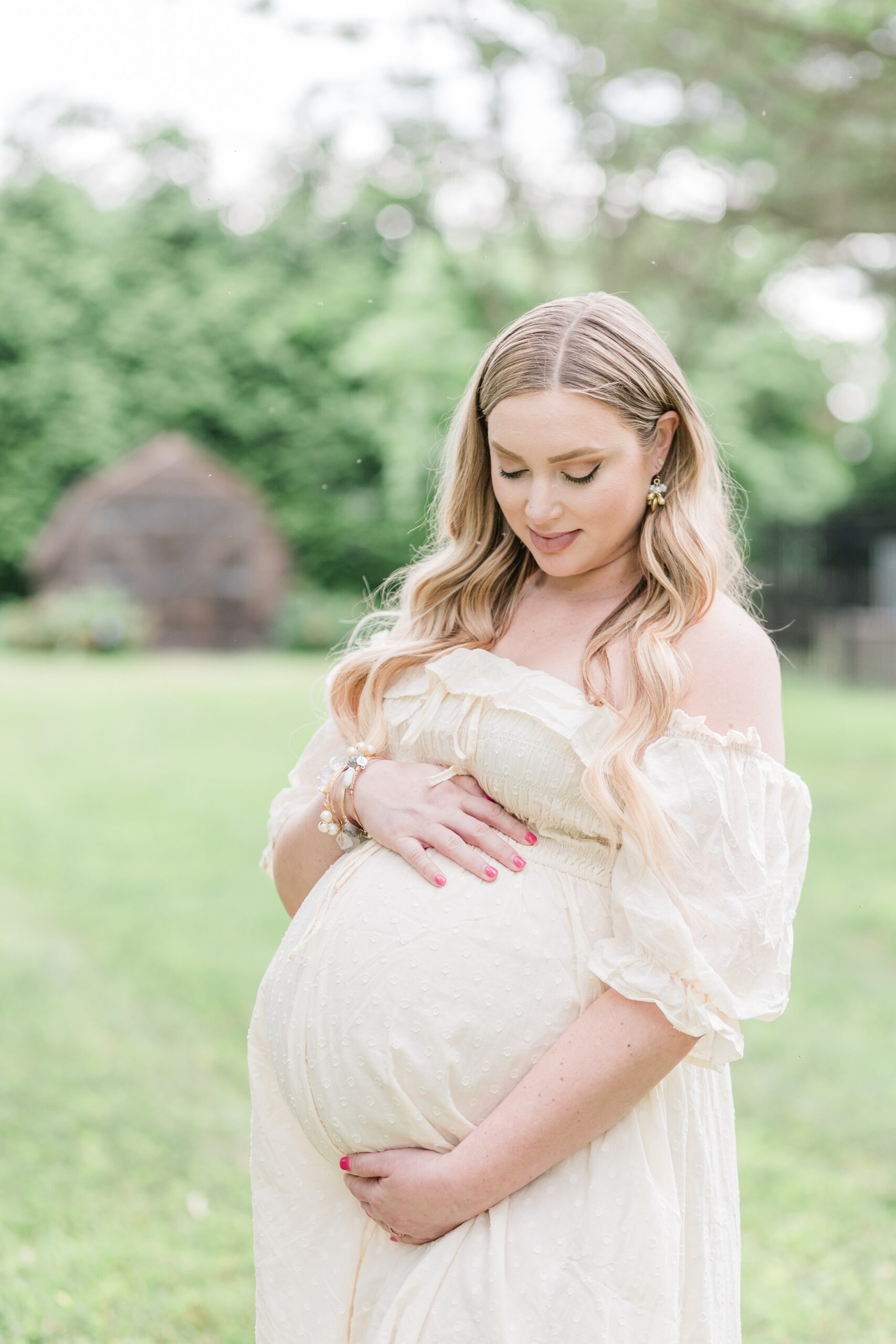 Mother in a soft yellow dress, cradles her twin pregnancy during a spring maternity session in her Gaithersburg, Maryland backyard, captured by a Maryland maternity photographer.