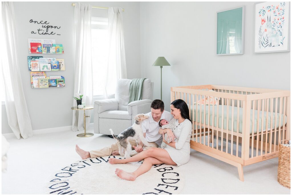 A family sits on the nursery floor with their newborn and dog, surrounded by soft natural light, neutral decor, and a simple wood crib—showcasing how to prepare a photo-ready space for an in-home newborn session.
