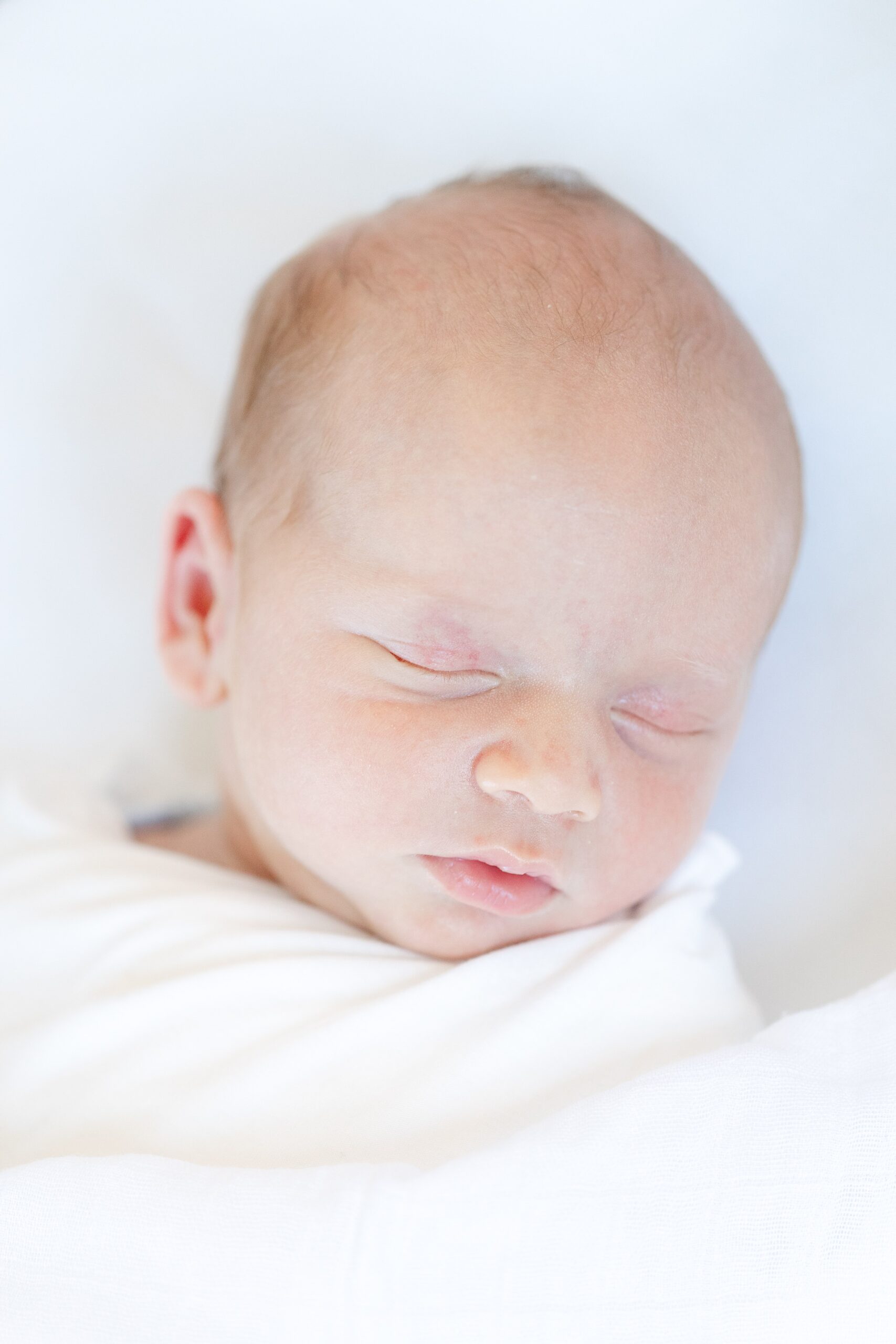 Close-up of a sleeping newborn swaddled in a soft white wrap, photographed in natural light—highlighting the simplicity and timeless styling recommended for in-home newborn sessions.