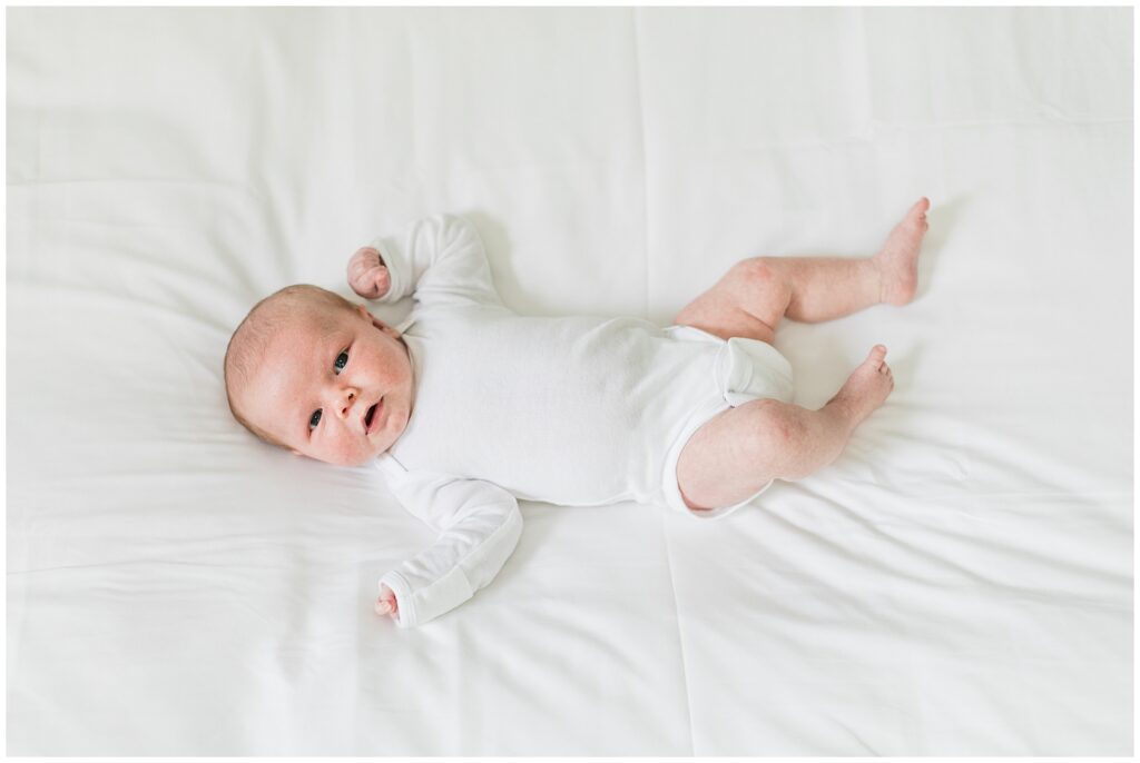 Newborn baby in a plain white long-sleeve onesie lying on a white bed, demonstrating a timeless and simple outfit for in-home newborn photography.