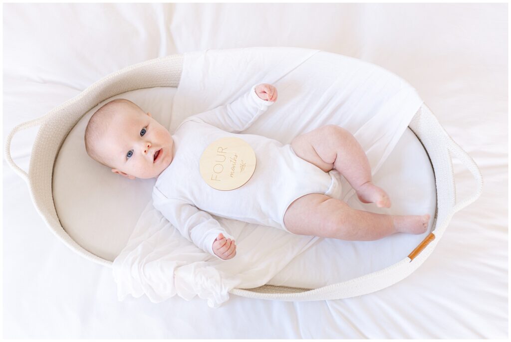 Four-month-old baby in neutral white outfit during lifestyle milestone photo session on a bed with a white bassinet and number four wooden sign