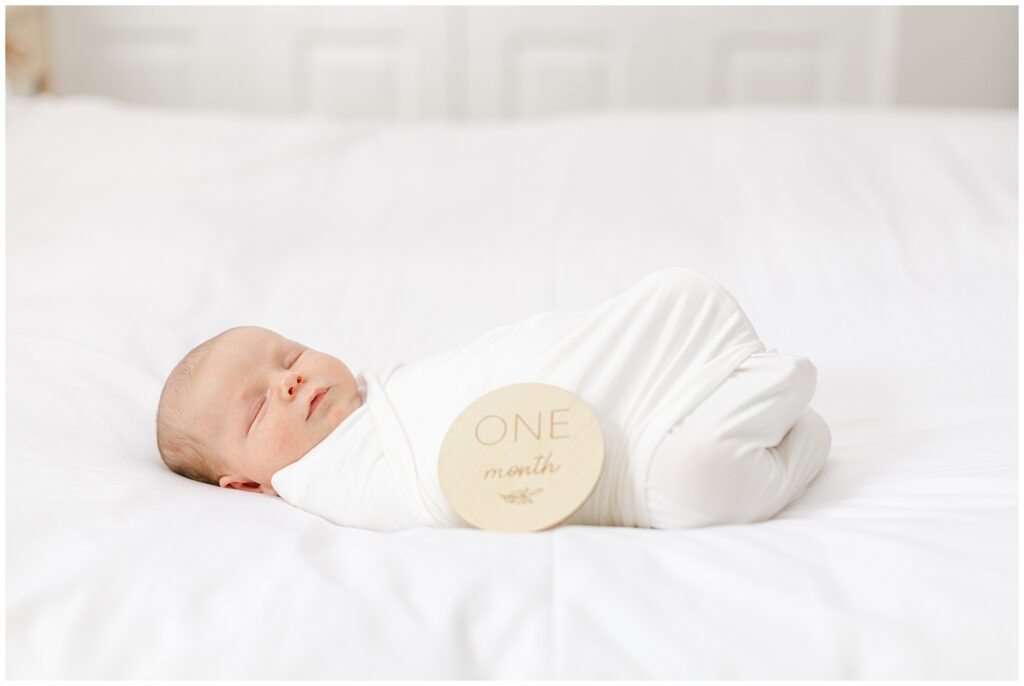 Baby in white onesie with neutral styling for their one-month milestone portrait on a bed in Howard County