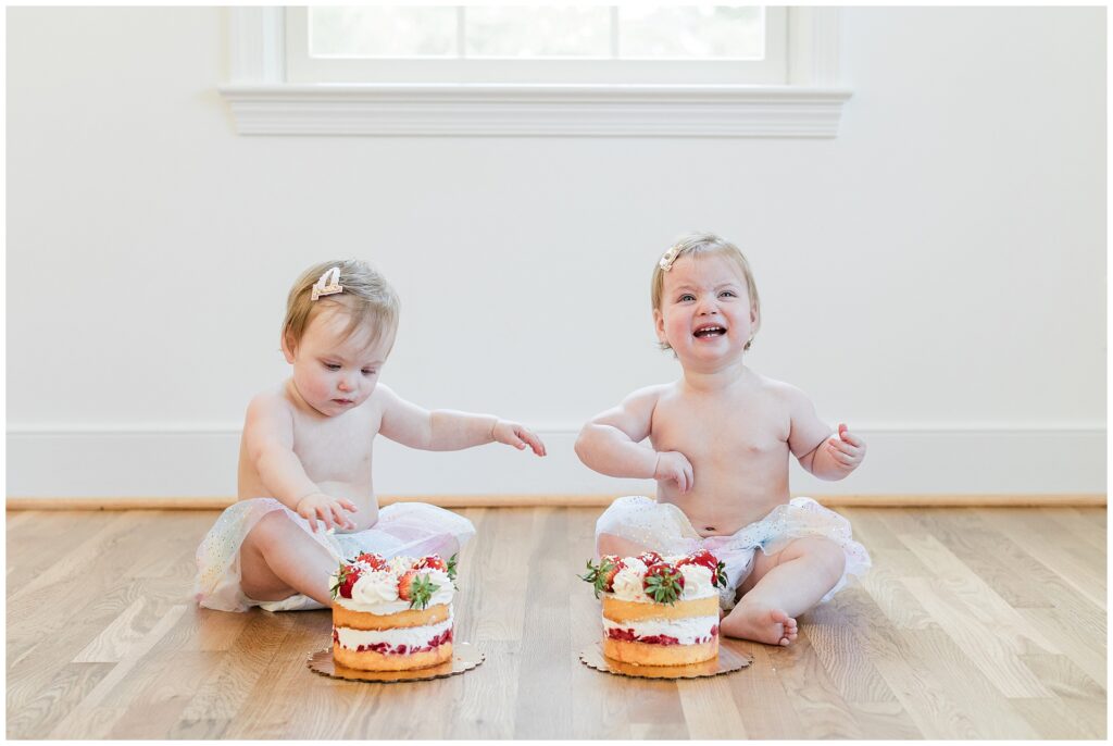 Twin girls sitting with individual strawberry shortcakes during a first birthday cake smash session at home in Gaithersburg, Maryland.