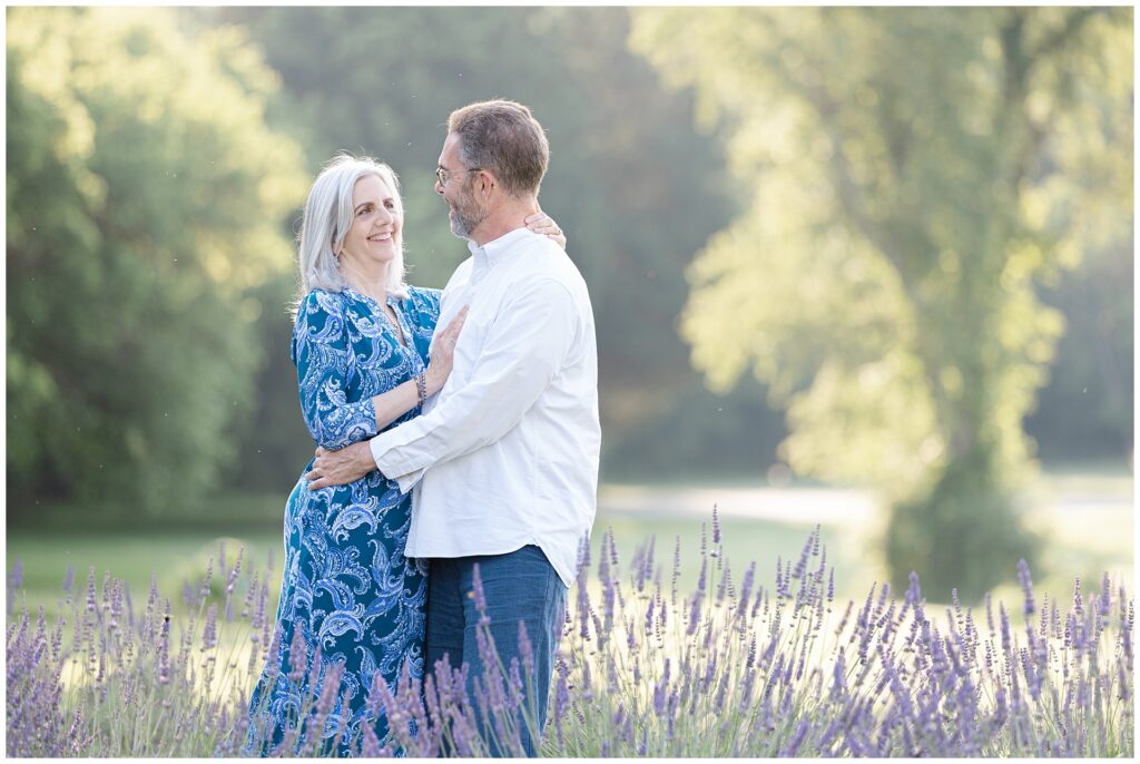 Newlywed couple in golden hour light during Gaithersburg lavender mini session, standing close in front of lavender rows; her bold blue patterned dress and his classic white shirt are elegant outfit choices for summer anniversary portraits