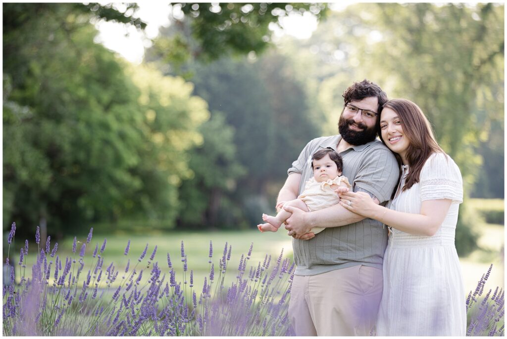 Parents holding four-month-old baby girl during Montgomery County mini session at Karis Lavender Farm; mother wearing short-sleeve white dress perfect for a summer lavender session with infants