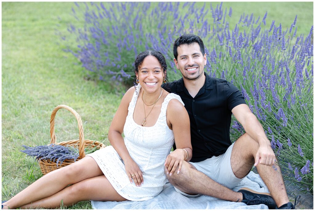 Young couple sitting in lavender field during Gaithersburg mini session, styled in light summer outfits; white eyelet dress with basket of lavender offers natural inspiration for what to wear to a lavender mini session in Maryland