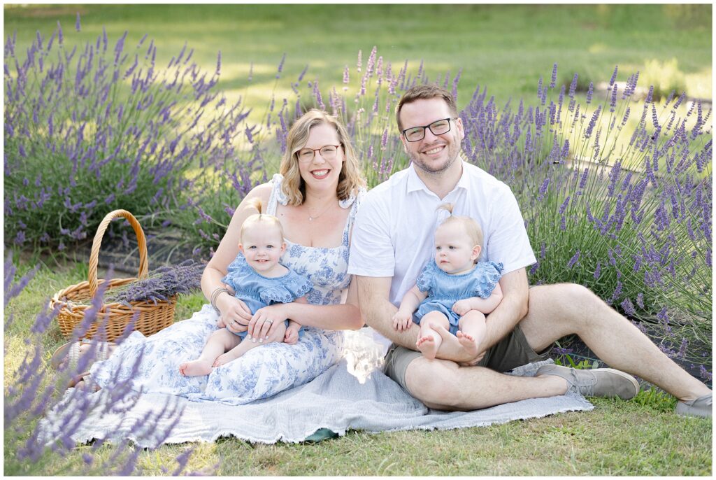Parents sitting in lavender field at Karis Lavender Farm with one-year-old twin girls during Montgomery County mini session; blue toile dress and matching baby rompers offer timeless outfit ideas for lavender family portraits