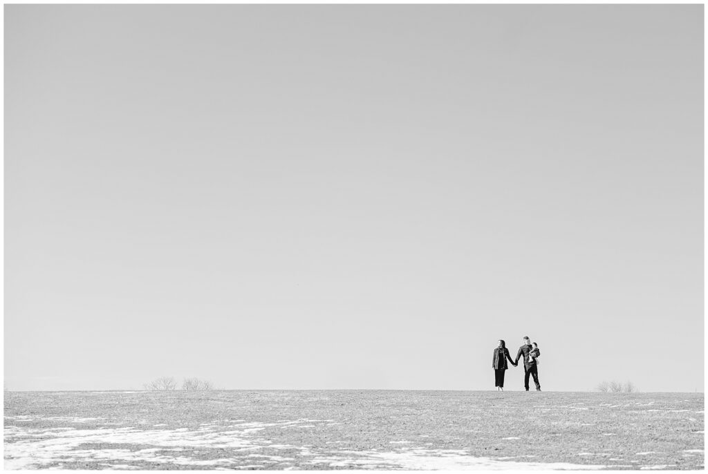 Dramatic black and white of a family of 3 on a hill with lingering snow. They stand hand in hand at Gaver Farm