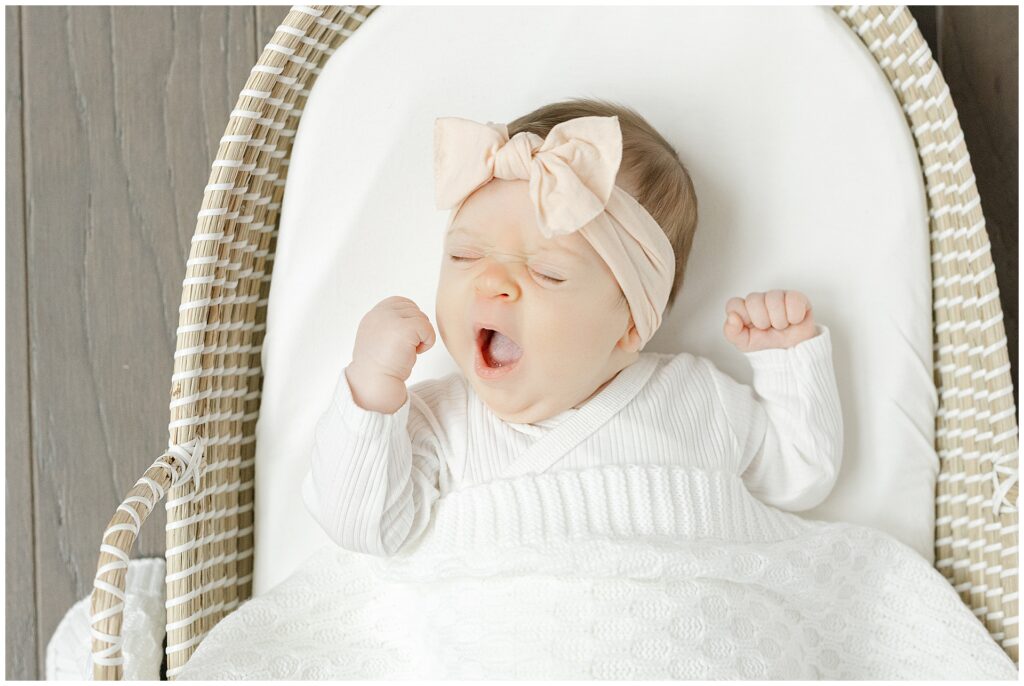 Baby girl in all white with soft pink headband, laying on a woven bassinet, yawns during her Arlington Virginia newborn session.
