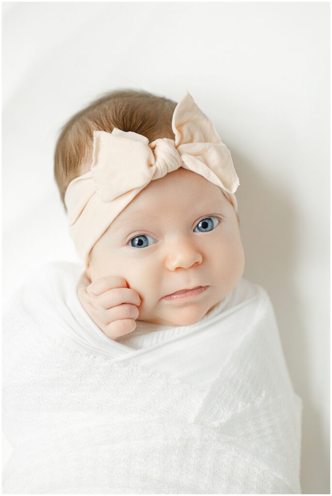 Baby girl in white swaddle with pink headband looks at the camera during her newborn session in Northern Virginia. Her bright blue eyes are alert and expressive.