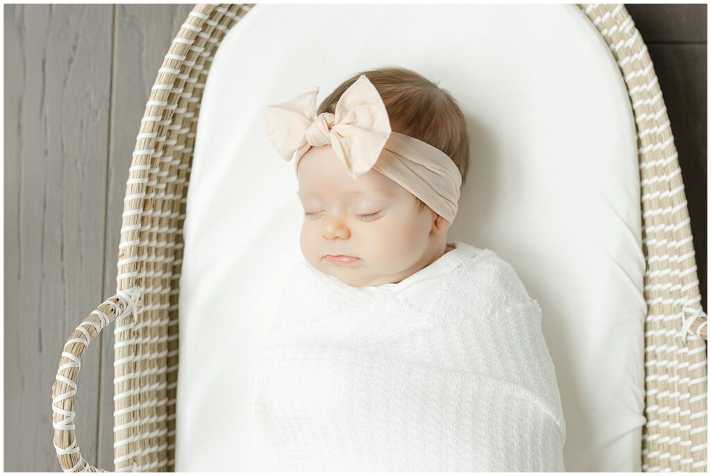 Newborn baby girl in a white swaddle and soft pink headband sleeps peacefully in a woven bassinet during her Northern Virginian Newborn session