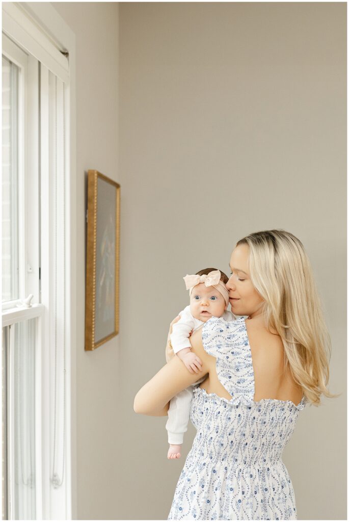Mother in a white and blue floral dress holds her baby girl in their newborn home session. Her lips barely brush against babys head in their Virginia newborn session