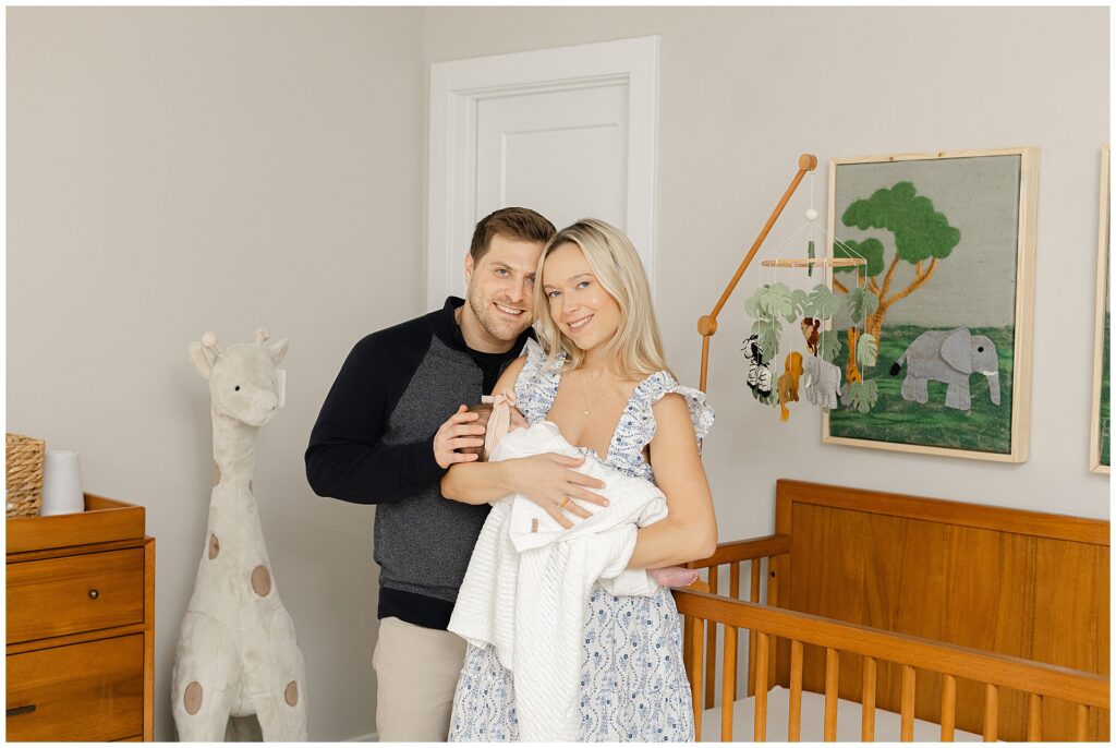 Mom in a white and blue floral dress  holds her baby girl in their newborn home session, in Virginia. Dad in a blue sweater hold his wife and they all smile at the camera.