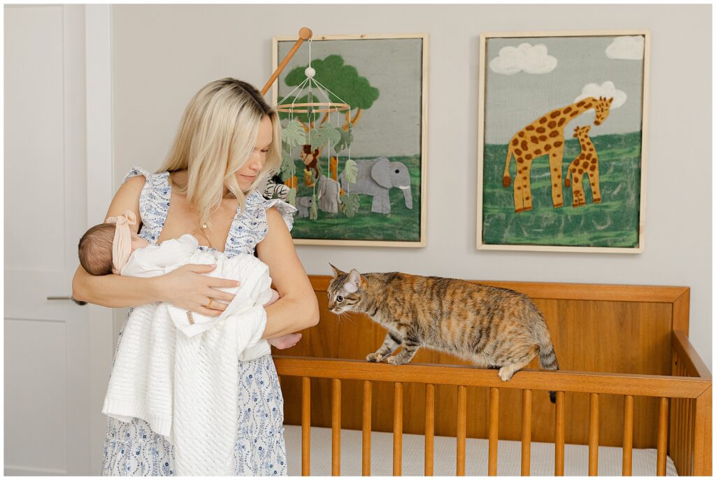 Mom in a white and blue floral dress  holds her baby girl in their newborn home session, in Virginia. Her cat balances on the crib