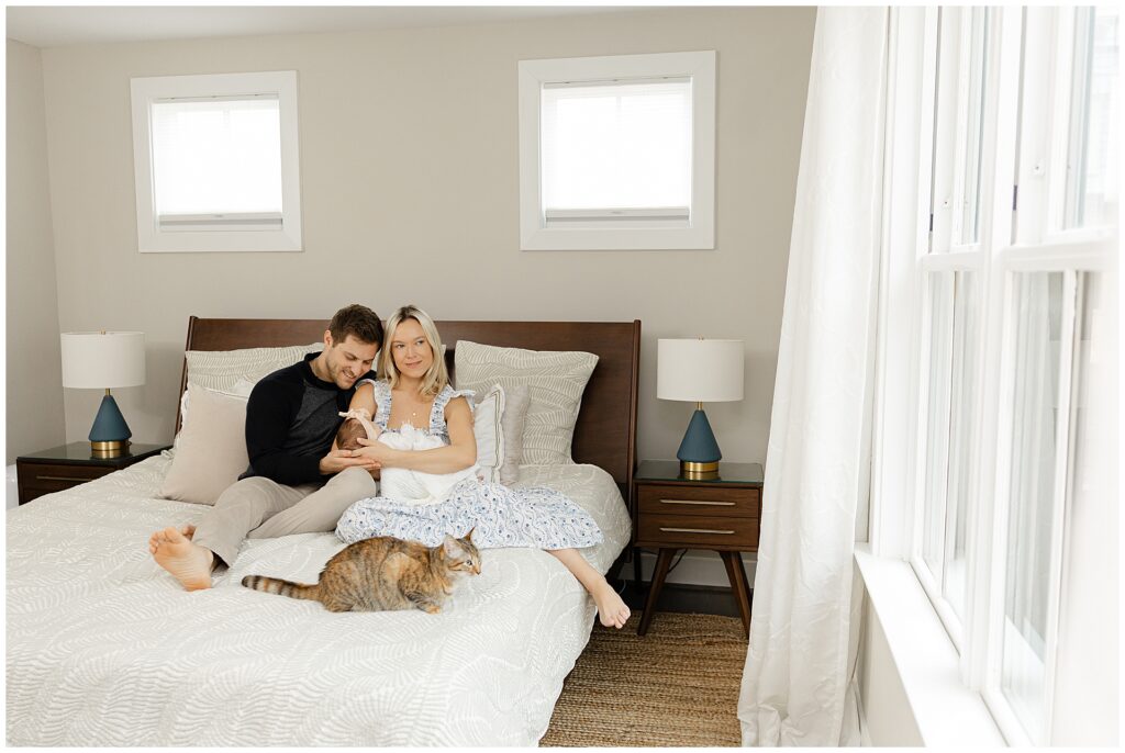 Mom in a white and blue floral dress  holds her baby girl in their newborn home session, in Virginia. Her husband cuddles with her on their bed with their cat in the foreground