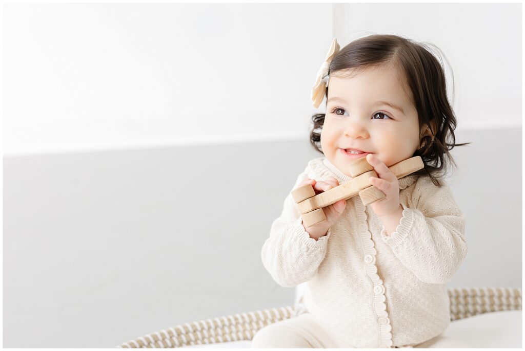 Close-up portrait of a one-year-old girl in a soft oatmeal sweater and hair bow hugging a wooden toy during first birthday photos in Annapolis