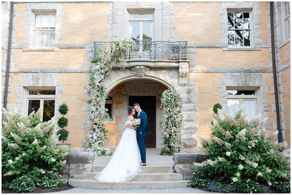 Wedding couple posing at the main entrance of Cairnwood Estate in Bryn Athyn, Pennsylvania, framed by a dramatic floral installation climbing the columns of the historic estate, showcasing a classic Cairnwood Estate wedding venue with grand architecture and romantic design details near Philadelphia.