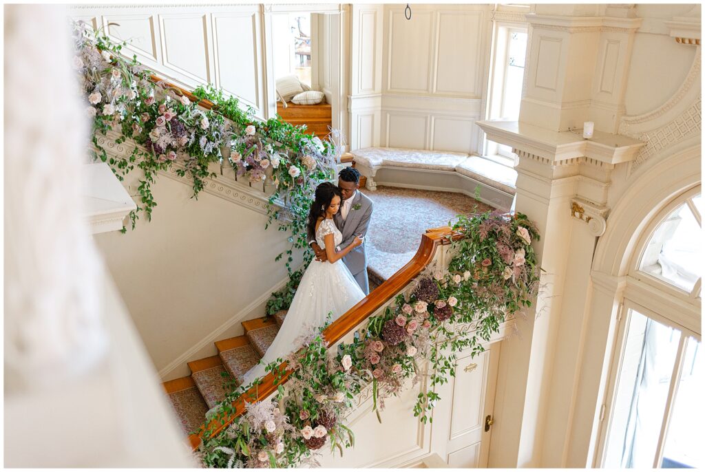 African American wedding couple posing together on the grand staircase inside the Great Hall at Cairnwood Estate, surrounded by soft mauve and lavender florals with cream roses that complement the historic architecture. A timeless portrait showcasing a Cairnwood Estate wedding venue ideal for romantic, refined celebrations at a Pennsylvania estate near Philadelphia.