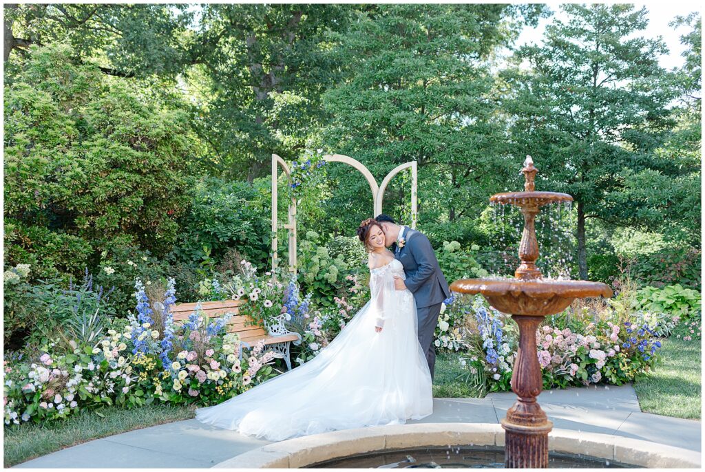 Wide view of the gardens at Cairnwood Estate in Bryn Athyn, Pennsylvania, with the historic fountain in the foreground and a wedding couple framed behind it, illustrating an intimate cocktail hour or portrait location at a Pennsylvania estate wedding venue near Philadelphia. This image highlights the romantic garden setting often used for relaxed wedding moments and guest mingling.