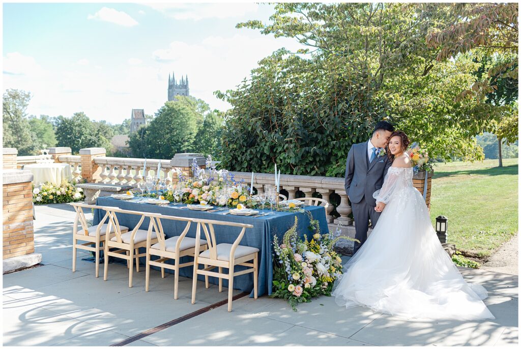 Wide view of a wedding couple on the South Terrace at Cairnwood Estate in Bryn Athyn, Pennsylvania, posing beside a reception table styled with wishbone chairs, a blue velvet tablecloth, and floral arrangements in blue, yellow, and orange, with Bryn Athyn Cathedral visible in the background. This image showcases a tented terrace reception option for a Cairnwood Estate wedding with scenic cathedral views near Philadelphia.