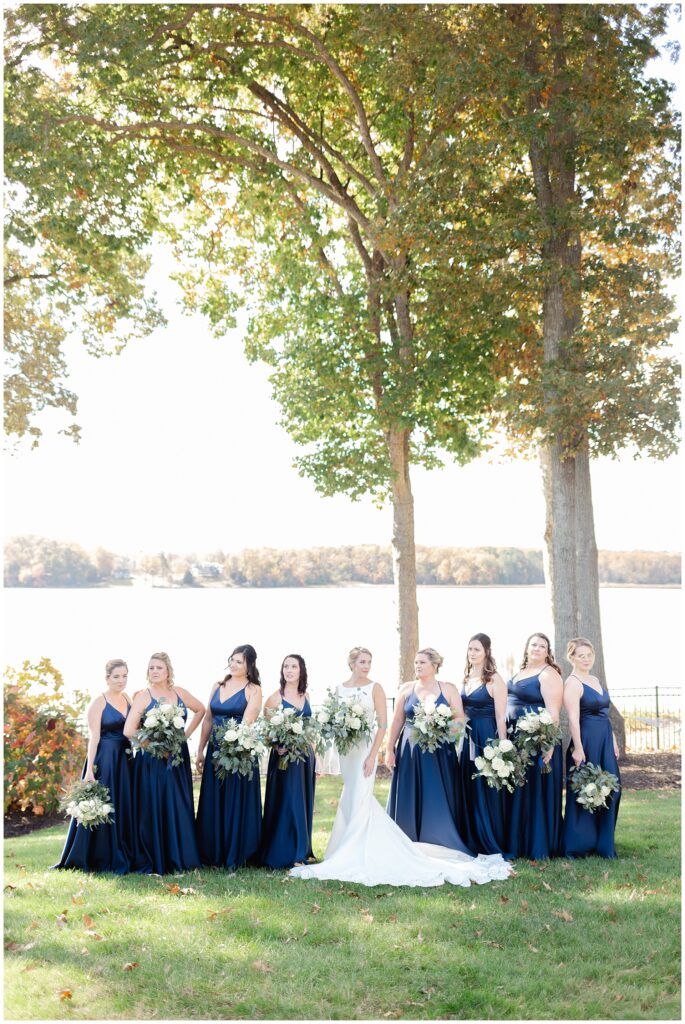 bride and bridesmaids pose in front of a tree, overlooking the potomac river during their fall waterfront wedding in maryland