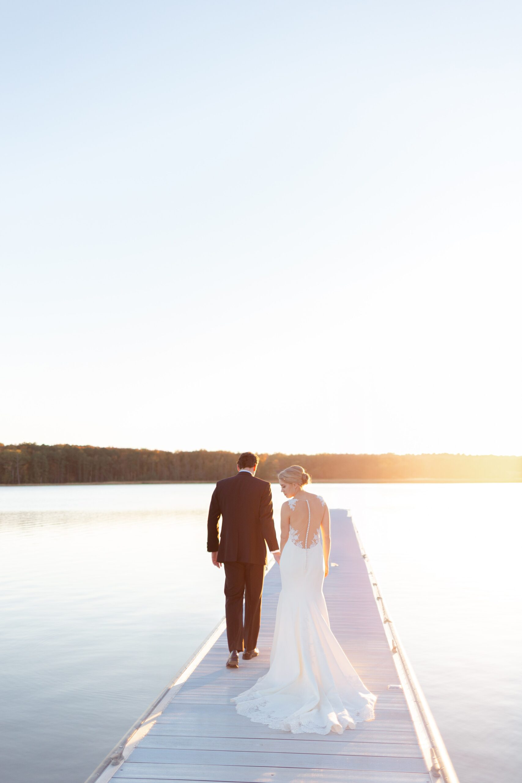 A bride and groom walk hand in hand down a pier, surrounded by the perfectly still water of the potomac river at their Maryland waterfront wedding. The sun flares to their right