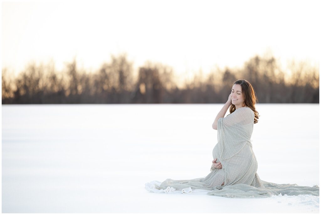 Fine art maternity photo of a woman in a silver dress, cradling her bump as she sits in her snow maternity session in maryland