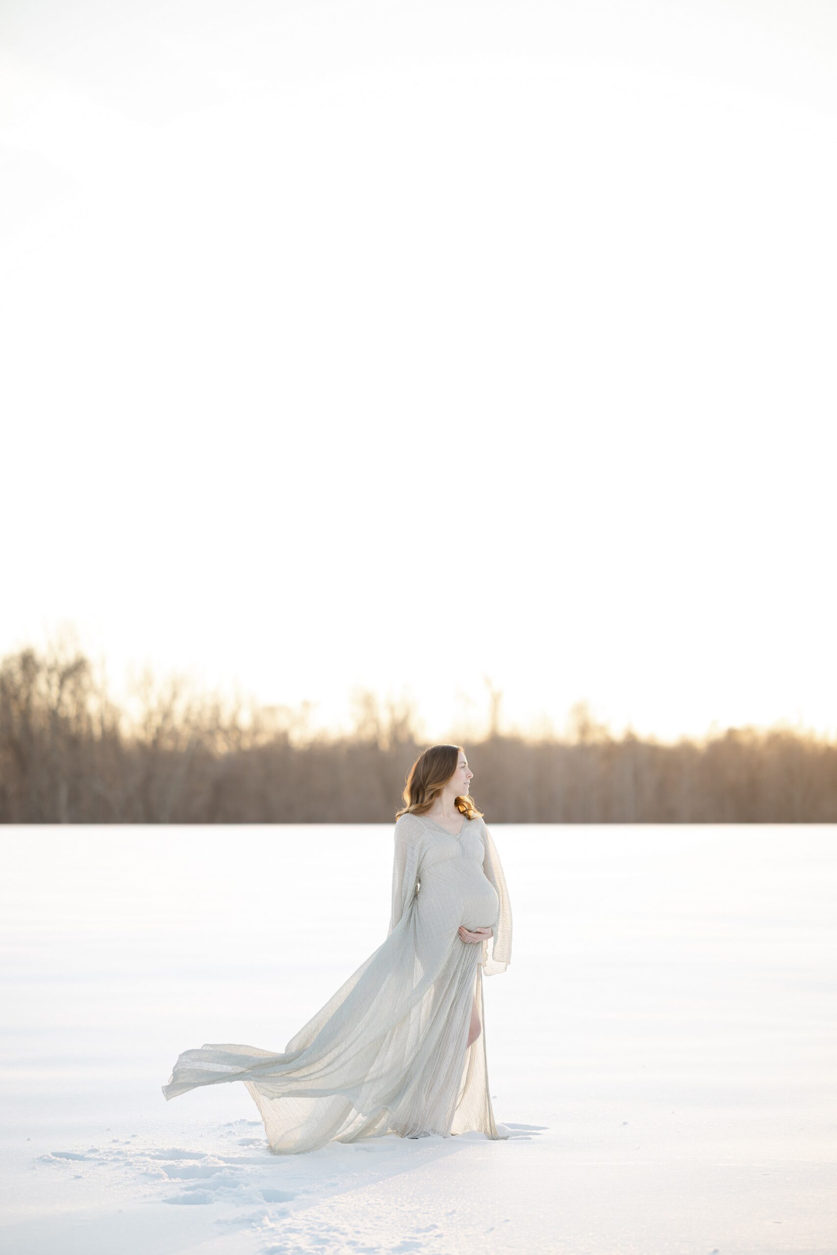 Pregnant woman standing in the snow looking off to the sunset as the wind tosses her silver dress during her snow maternity session in Maryland