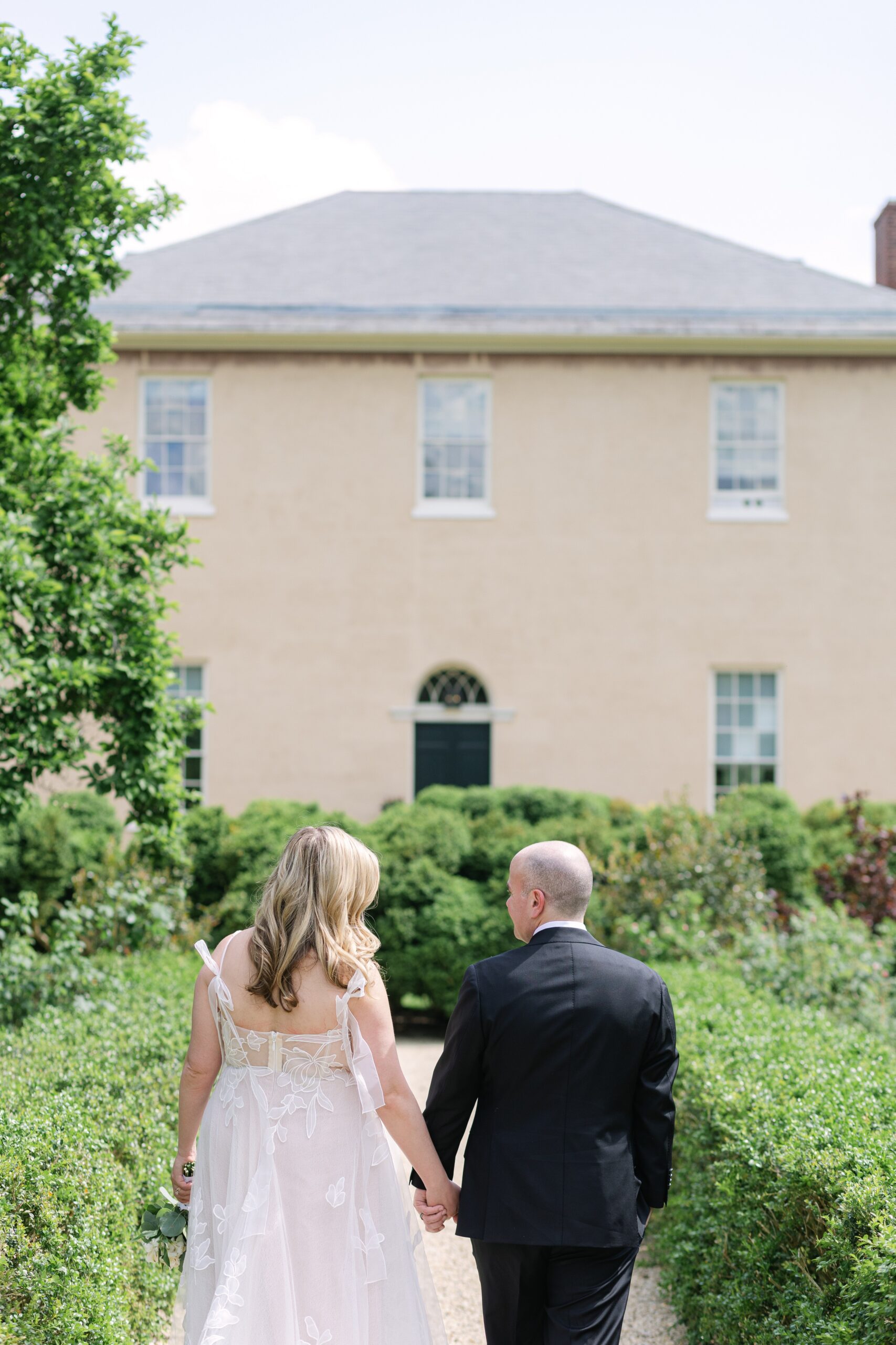 Bride and groom walking hand in hand away from the viewer, through the gravel path at the Tudor Place during their Wedding