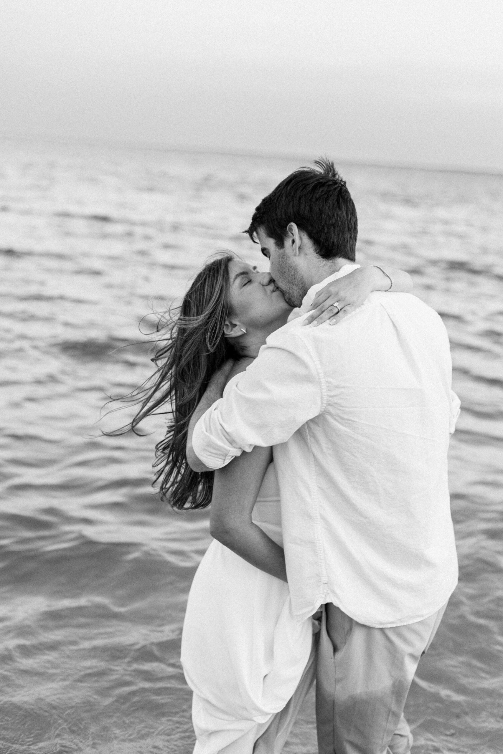Cinematic black and white engagement session at a maryland beach. Couple in in white kisses while the wind blows their hair around while they stand in the water