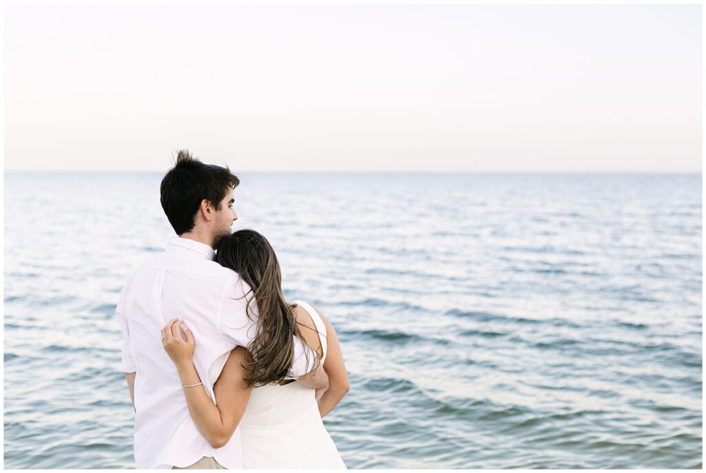Moody blue hour beach engagement photos in Maryland. Couple hugs as they  look at the twilight over the Chesapeake during a Maryland beach engagement session.